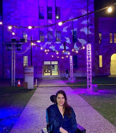 A young woman in a wheelchair in UQ's Great Court at night, with buildings lit up purple.