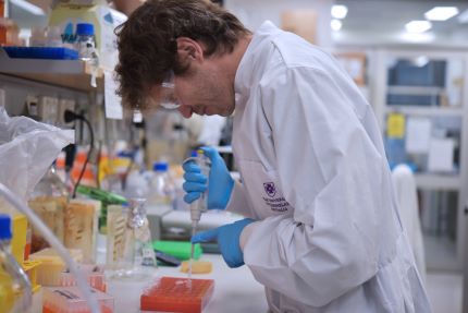 A male scientist uses a pipette at a lab bench. He's wearing a lab coat, goggles and gloves.