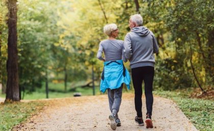 two people with grey hair are walking away from the camera along a sandy track with green vegetation either side