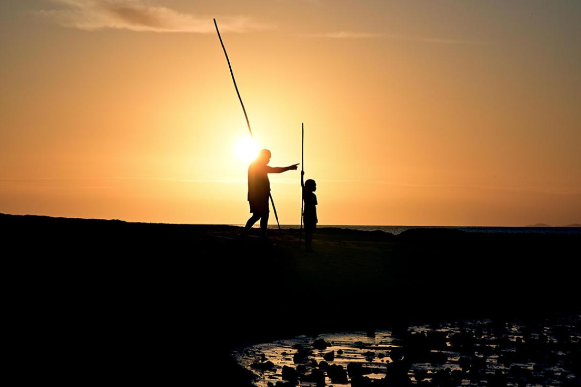 Two people holding spears standing by a lake, the sun setting behind them 