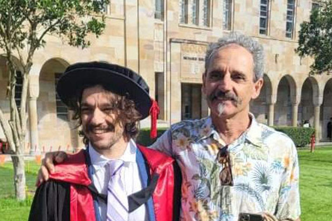 A young man in a graduation gown standing with his Father in front of a sandstone building 